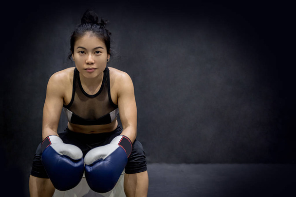 Hermosa joven asiática feliz mujer sentada y posando con guantes de boxeo azul en el gimnasio, fondo negro
 - Foto, Imagen