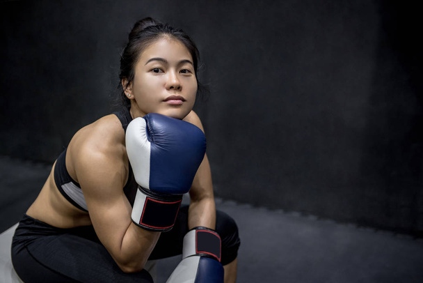 Belle jeune femme asiatique heureuse assise et posant avec des gants de boxe bleus dans la salle de gym, fond noir
 - Photo, image