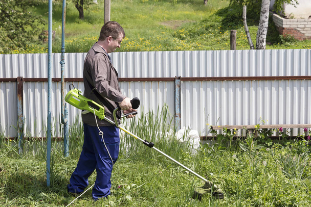 un hombre en vasos con un trimer corta la hierba en el patio de una casa en el pueblo, pone orden en el territorio del país
 - Foto, Imagen