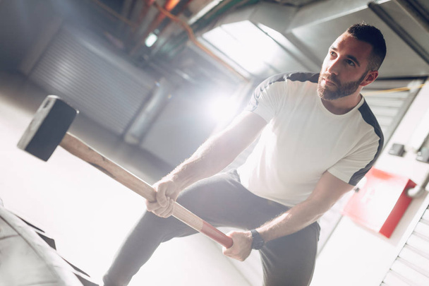 Молодой мускулистый человек бьет молотком по огромной шине на тренировках в гараже
 - Фото, изображение