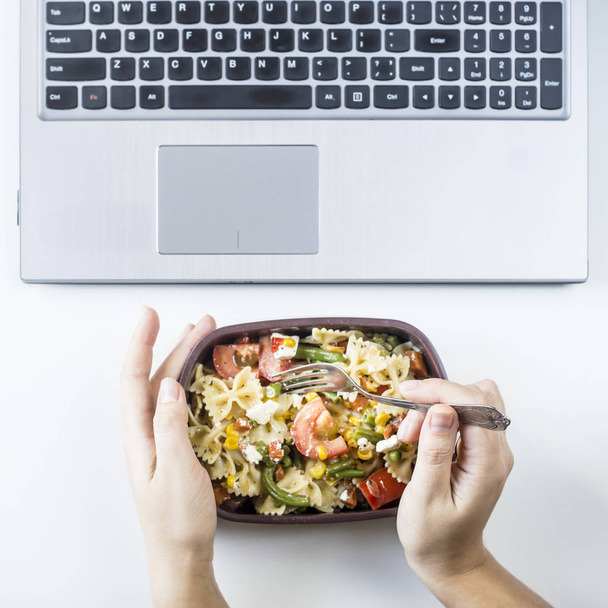 Container with salad with pasta in the workplace near the computer. Lunch in the office during a break between work. Woman take a food. Top view, flat lay - Fotó, kép