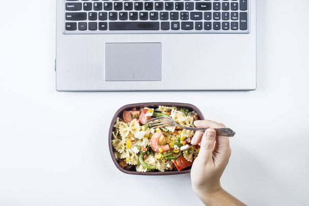 Container mit Salat mit Pasta am Arbeitsplatz in der Nähe des Computers. Mittagessen im Büro während einer Arbeitspause. Frau nimmt Essen. Draufsicht, flache Lage - Foto, Bild