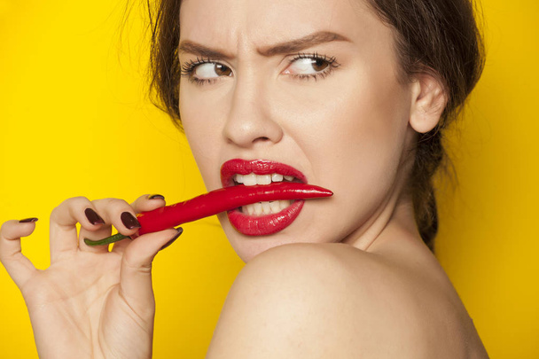 Mladá sexy žena kousání chili pepper na žlutém podkladu - Fotografie, Obrázek