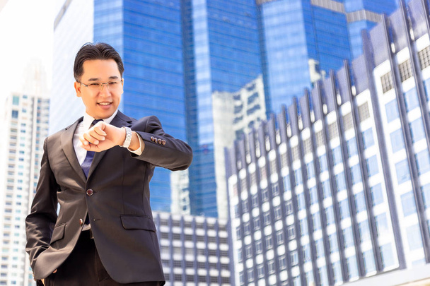 Handsome businessman is looking wristwatch for checking time. Charming handsome guy gets appointment and he comes to meeting point early. He feels relieved. He wear full suit. high building background - Photo, Image