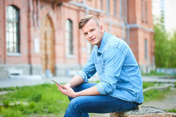 студент в джинсовой рубашке с планшетным ПК сидит рядом с университетом в солнечный день
  - Фото, изображение