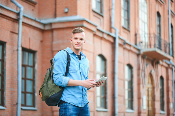 student in denim shirt holding tablet pc in hands and looking at camera while standing near university  - Фото, изображение