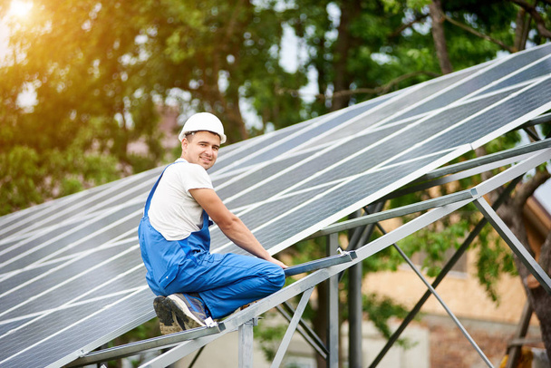 Joven trabajador electricista sonriente sentado en casi terminado sistema de panel fotovoltaico solar independiente sobre fondo de árbol verde brillante. Concepto de energía alternativa
. - Foto, imagen