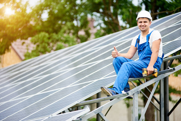 Joven electricista sonriente sentado en casi terminado sistema de panel solar fotovoltaico independiente con destornillador, mostrando gesto pulgar hacia arriba sobre fondo de árbol verde brillante
 - Foto, imagen