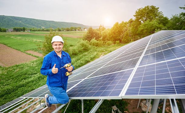 Construction worker with screwdriver looking in camera with thumb-up gesture on photo voltaic panel solar system shiny surface and lit by sun green fields background. Alternative energy concept. - Photo, Image
