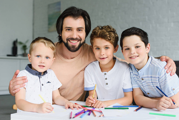 πορτρέτο του χαμογελώντας ο πατέρας και γιοι στο τραπέζι με έγγραφα και πολύχρωμα μολύβια στο σπίτι - Φωτογραφία, εικόνα