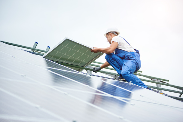Aurinkopaneelijärjestelmän asentaminen. Ammattimainen työntekijä kypärä ja sininen yleinen nosto aurinkopaneeli metalli alustalla. Vaihtoehtoinen energia- ja ammattirakentamisen käsite
. - Valokuva, kuva