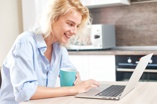 leuke vrouw met koffiekopje zittend aan tafel in de keuken bezig met laptop - Foto, afbeelding