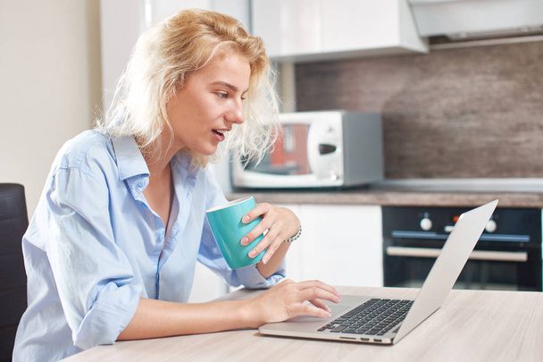 cute woman with coffee cup working on laptop while sitting at table in kitchen - Fotoğraf, Görsel