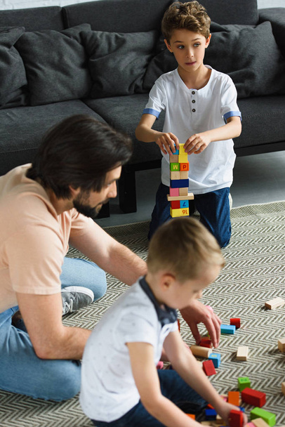 side view of father and kids playing with wooden blocks together on floor at home - Foto, afbeelding