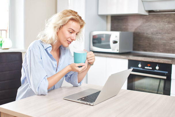 niedliche Frau hält Kaffeetasse und sitzt am Tisch mit Laptop in Küche, Frühstückskonzept  - Foto, Bild