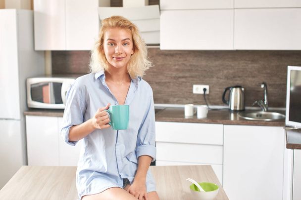beautiful blonde woman with blue teacup sitting on wooden table in kitchen - Fotografie, Obrázek