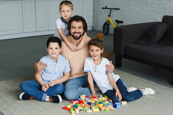 pai feliz abraçando filhos pequenos e olhando para a câmera enquanto sentado no chão com blocos de madeira em casa
 - Foto, Imagem