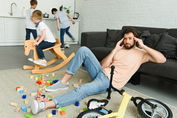 padre estresado sentado en el suelo y los niños jugando en la habitación
 - Foto, Imagen