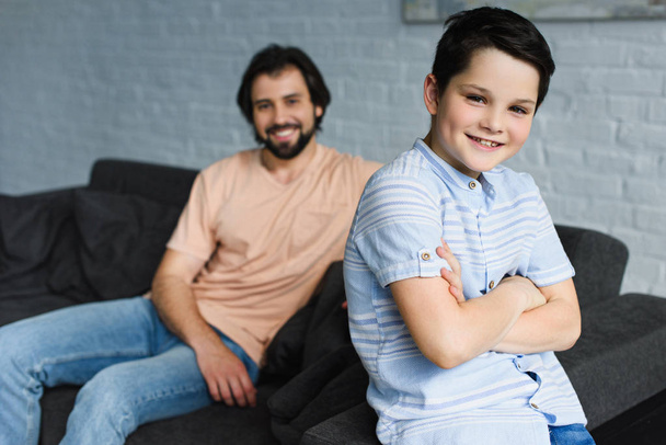 selective focus of smiling boy with arms crossed and father resting on sofa behind at home - Fotoğraf, Görsel