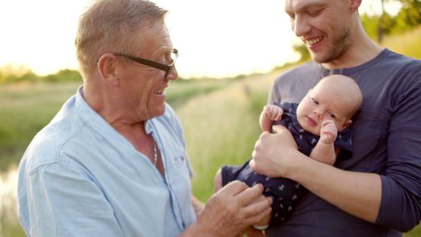 Multi-generation family. Grandfather kisses the foot of a newborn grandson. Three men rest on the nature, near the river. Colorful sunset - Footage, Video