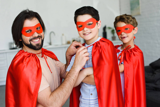 portrait of smiling father and sons in red superhero costumes looking at camera at home - Foto, imagen