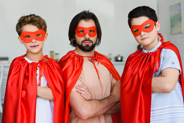 Porträt von Vater und Söhnen in roten Superheldenkostümen mit verschränkten Armen, die zu Hause in die Kamera schauen - Foto, Bild