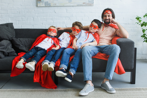 smiling father sitting near sleeping sons in red superhero costumes on sofa at home - Foto, Imagen