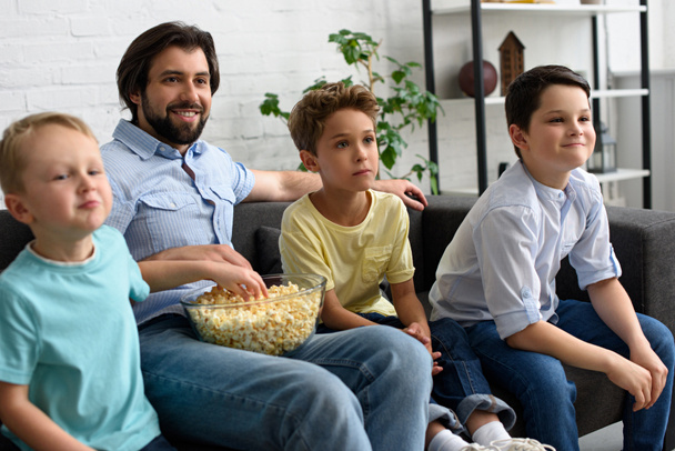 smiling man and little sons with popcorn watching film together at home - Photo, image