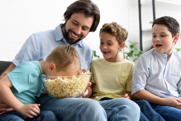 smiling man and little sons eating popcorn while watching film together at home - Foto, afbeelding