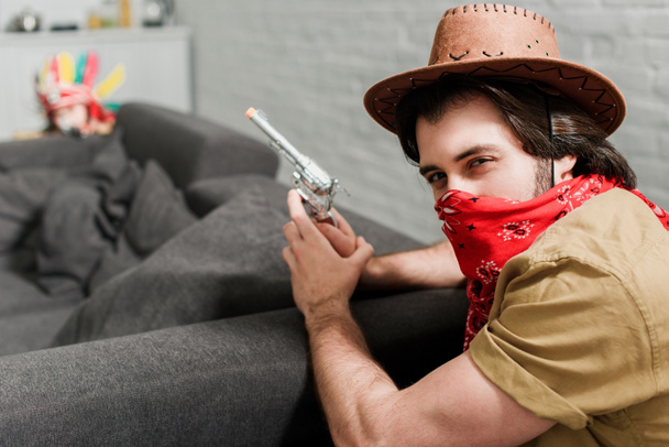 side view of man in red bandana and cowboy hat with toy gun at sofa at home - Photo, Image