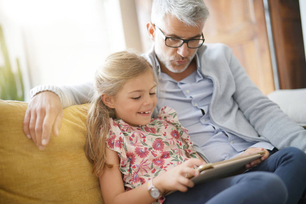 Father and daughter connected with tablet, sitting on couch - Photo, Image