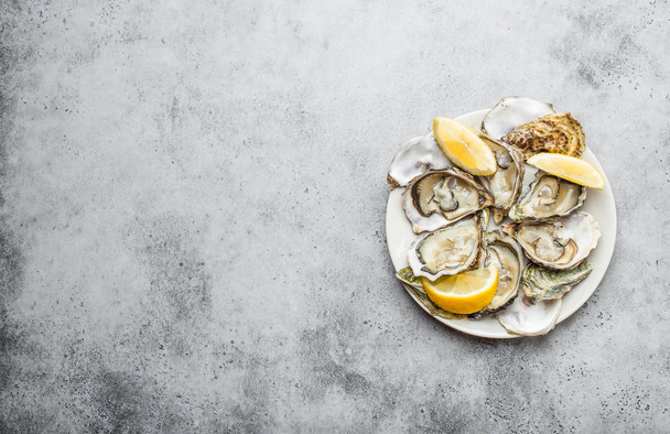 Close-up de meia dúzia de ostras abertas frescas e conchas com cunhas de limão em um prato, vista superior, fundo de concreto rústico cinza, espaço para texto. Ostras delicadas para lanche, almoço
 - Foto, Imagem