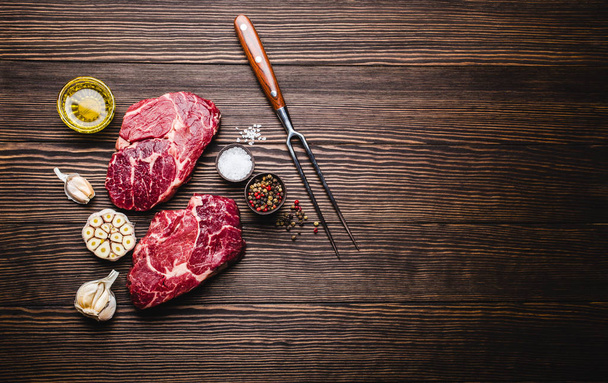 Raw marbled meat steak Ribeye on rustic wooden background with fork, seasonings and olive oil for cooking, roasting, grill or BBQ party with space for text, top view. Meat restaurant concept - Фото, изображение