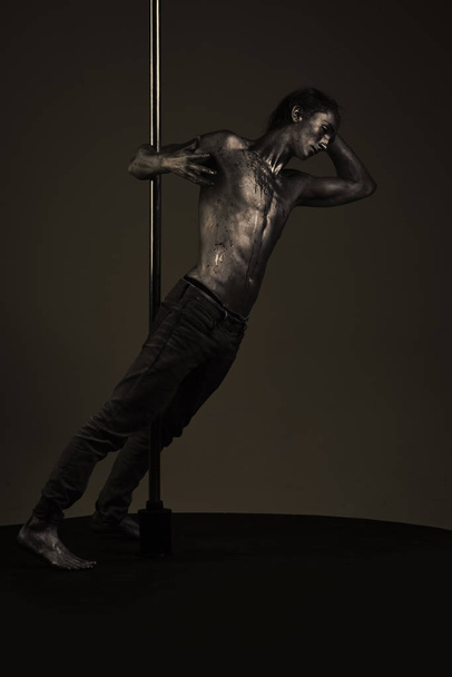 Pole dance. Man with nude torso covered with shimmering paint, dark background. - Фото, изображение