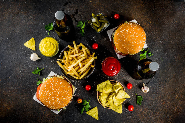 Various party food, Hamburgers, French fries, potato chips, pickled cucumbers, onions, tomatoes and cold beer bottles, rusty black concrete background copy space - Photo, Image