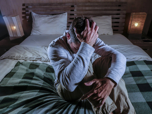 Porträt eines jungen verzweifelten und depressiven Mannes zu Hause im Schlafzimmer, der traurig und verwirrt auf dem Bett sitzt und Schmerzen und Depressionen leidet und weint. - Foto, Bild