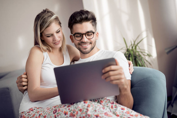 Hermosa pareja joven usando el ordenador portátil y sonriendo mientras se sienta en el sofá en casa
. - Foto, Imagen