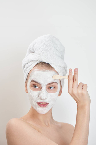 smiling woman wearing towel on head applying face mask with wooden stick , beauty treatment concept  - Photo, Image