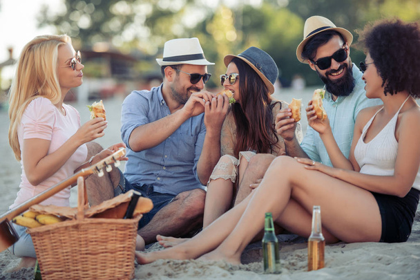 Ομάδα νέων που τρώνε σάντουιτς κατά τη διάρκεια πικ-νικ στην παραλία. - Φωτογραφία, εικόνα