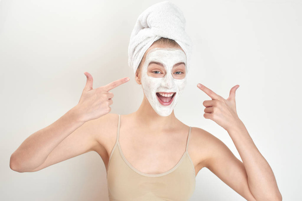 beautiful woman with towel on head pointing by fingers at mask on face, beauty treatment concept  - 写真・画像