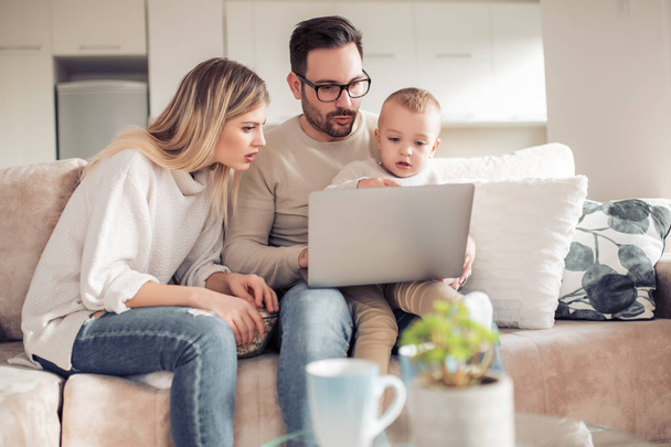 Gyönyörű fiatal család használ laptop, beszél, és mosolyogva, miközben ül a kanapén, otthon. - Fotó, kép