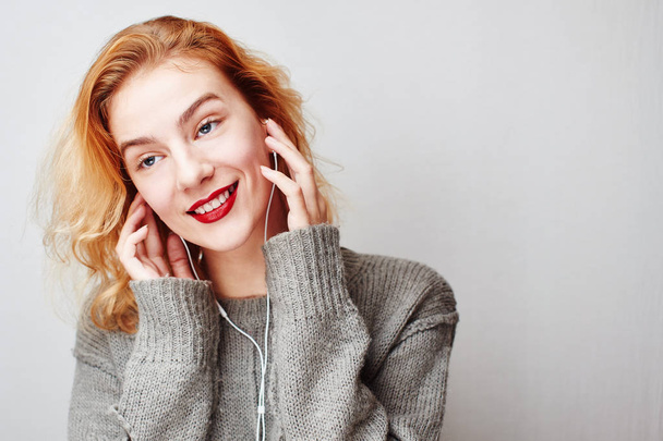 portrait of young red-haired woman in sweater listening to music with headphones on gray background  - Fotó, kép