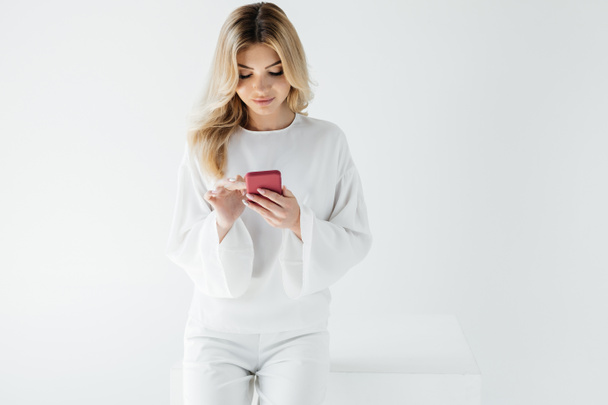 portrait of blond woman in white clothing using smartphone isolated on grey - Fotografie, Obrázek