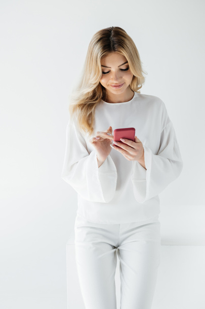 smiling blond woman in white clothing using smartphone isolated on grey - Foto, Imagen