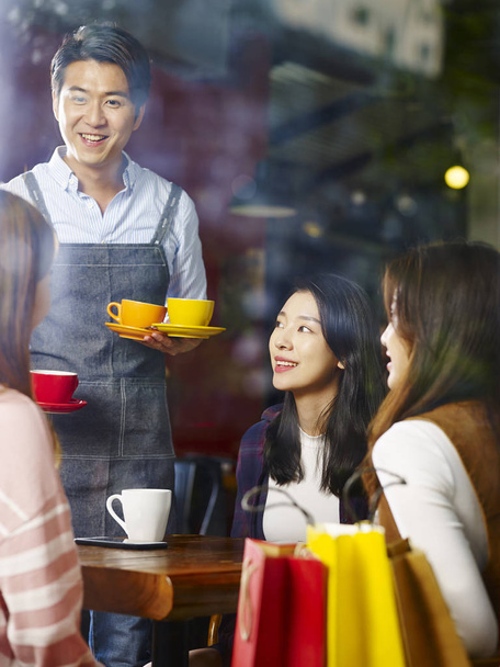 молодий щасливий усміхнений азіатський офіціант, що обслуговує жінок-клієнтів у кав'ярні, знятий через віконне скло
. - Фото, зображення