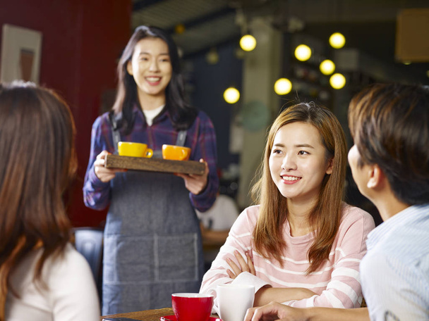 チャットのテーブルに座っている 3 幸せな若いアジア大人サービングのコーヒー、ウェイトレスしながら話している前景の女の子に焦点を当てる. - 写真・画像