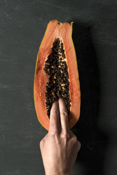 high angle shot of the the fingers of a young caucasian man into a papaya, cut in half, on a dark green background - Photo, Image