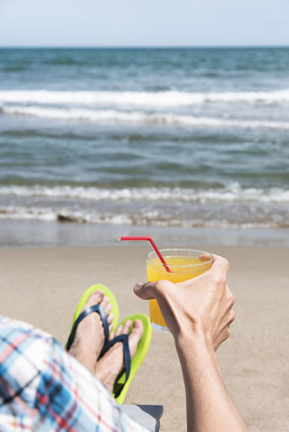 closeup of a young caucasian man relaxing on the beach, sitting on a deck chair and wearing a pair of green flip-flops, with a glass with a refreshing orange drink in his hands - Фото, изображение