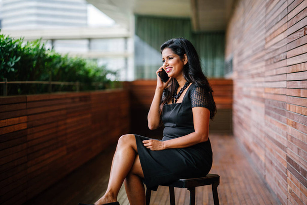 Portrait of a Indian Asian business woman speaking on her smartphone. She looks concerned and worried as she talks on her phone and is sitting in a chair in a corridor with wooden walls in the day. - Фото, изображение