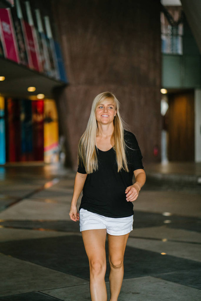 Portrait of a young and athletic blond woman walking in her urban campus in Asia during the day. She is dressed in comfortable, sporty and casual attire and sports shoes and she is smiling as she walk - Foto, afbeelding